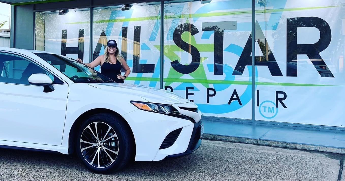 Woman standing beside a white car in front of Hail Star Repair shop with large sign.